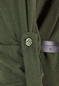 Groene stoffen mouw met knoopsluiting en gestructureerd weefsel naast een donkere leren ceintuur op een kledingstuk.