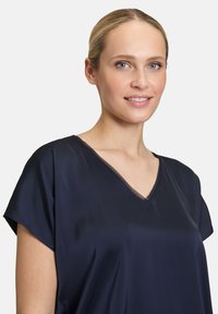 Top blu navy a maniche corte in tessuto liscio e lucido con scollatura a V e rifiniture trasparenti, caratterizzato da una vestibilità ampia e rilassata.