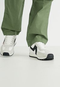 Weiße und graue Turnschuhe mit schwarzen Akzenten, ausgestattet mit einem strukturierten Obermaterial, Schnürung und einer Gummisohle, getragen mit grünen Hosen.