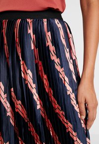 Närbild av en plisserad kjol med marinblå, rosa och röda geometriska mönster, bärs med en korallfärgad topp som visar höger arm och hand.