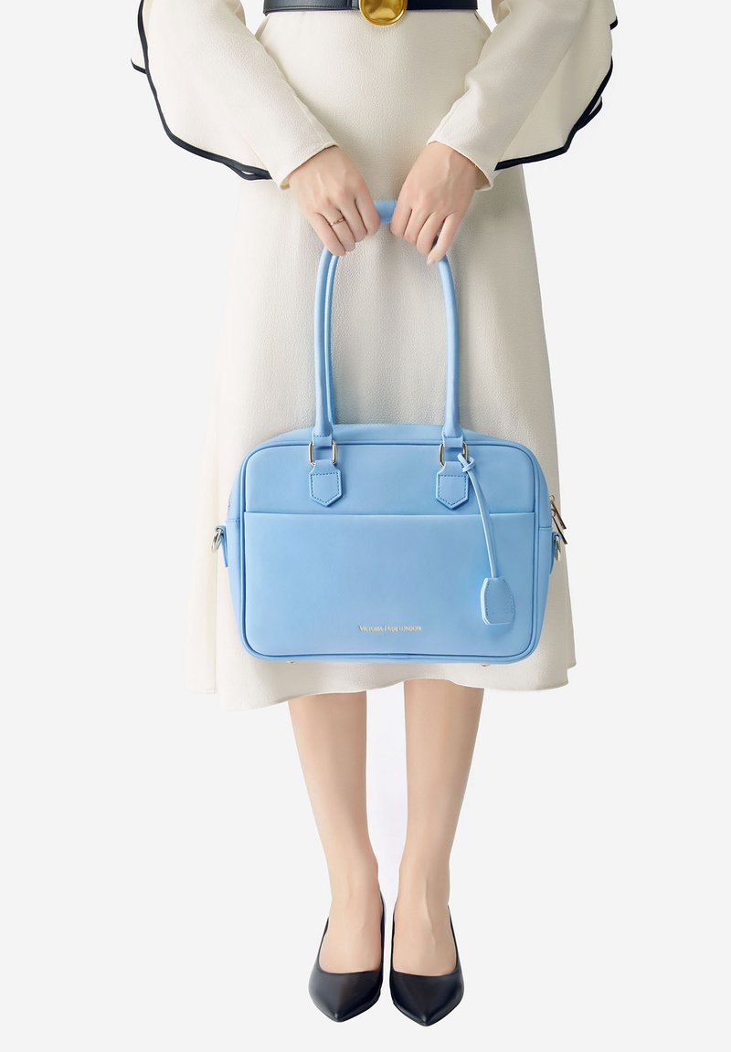 Bolso de cuero azul claro con dos asas, de forma rectangular, que presenta herrajes dorados y un bolsillo con cremallera. Superficie lisa con detalles de costura.