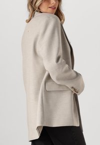 Blazer de color beige claro con textura suave, botón único en el puño, bolsillos laterales y un dobladillo trasero ligeramente más largo.