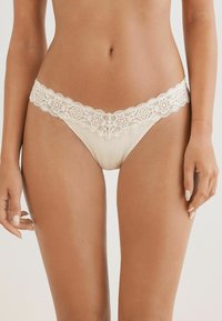 Beige kanten string met golvende randen, voorzien van ingewikkelde bloemenpatronen langs de tailleband en een gladde, zachte stof.