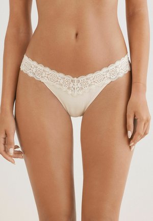 String en dentelle beige avec des bords festonnés, présentant des motifs floraux complexes le long de la taille et un corps en tissu lisse et doux.