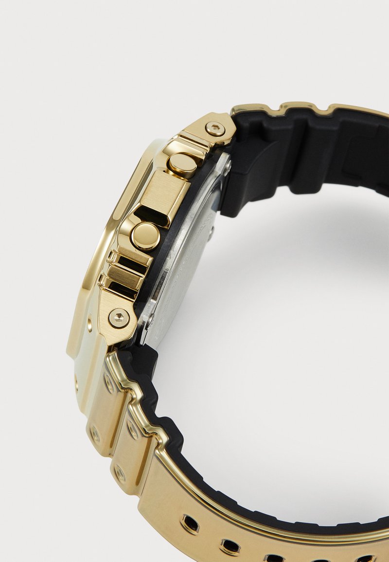 Orologio da polso in metallo dorato con cinturino in gomma nera. Caratteristiche includono pulsanti e una fascia testurizzata per una presa migliore. Design minimalista e durevole.