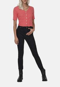 Cardigan court corail à manches courtes et avec un bouton devant, associé à un jean noir taille haute et des bottines noires. Texture de tissu lisse.