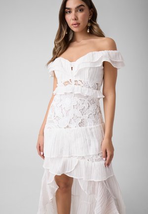 Mujer con un vestido blanco sin hombros con detalles de encaje y volantes, de pie frente a un fondo liso gris claro.