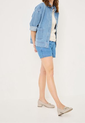 Vrouw in een lichtblauwe spijkerjas en shorts met een crème sweater en beige lage instappers, staand tegen een effen achtergrond.