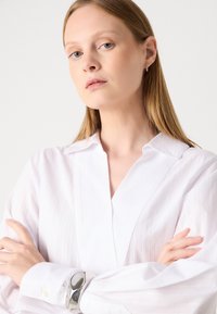 Hvit stripete bluse med spiss krage, lange ermer og knappedetaljer. Har en glatt tekstur og svak glans, komplementert av et sølvarmbånd.