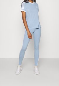 Žena v světle modrém tričku Adidas a legínách s bílými pruhy a bílými teniskami stojí na jednobarevném pozadí.