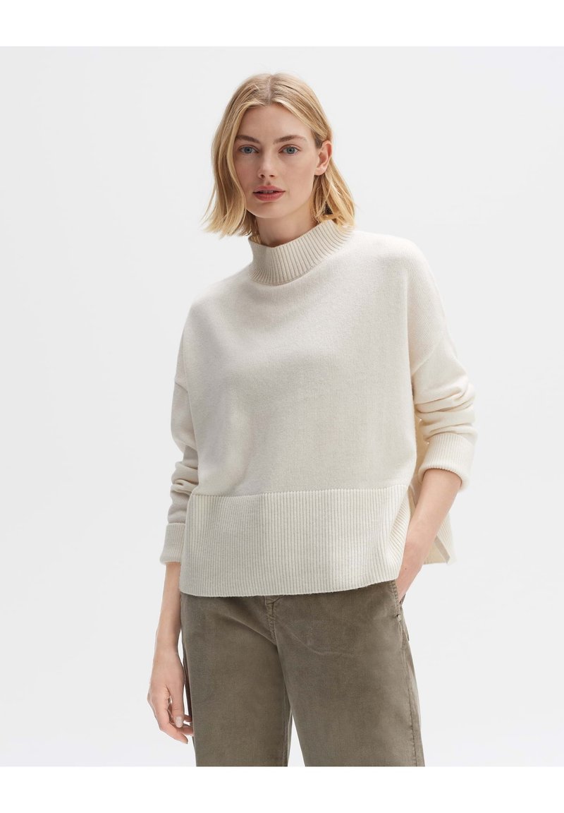 Cremfarbener Pullover mit hohem Kragen, geripptem Saum und Ärmeln. Weiche Textur, lockere Passform, kombiniert mit olivgrünen Hosen. Minimalistisches Design.