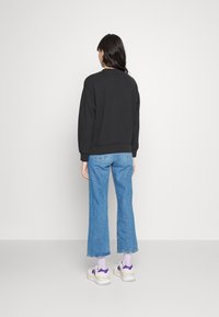 Czarny sweatshirt z okrągłym dekoltem, połączony z niebieskimi dżinsami o średnim stanie. Modelka ma na sobie masywne białe sneakersy z fioletowymi akcentami.