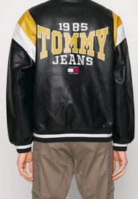 Musta nahkatakki, jossa keltaisia, valkoisia ja mustia yksityiskohtia. Takana näkyy näyttävä "TOMMY JEANS" -teksti ja "1985", resorireunukset hihoissa ja helmassa.