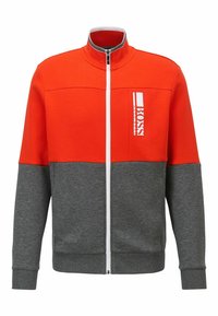 Mikina na zip s oranžovou horní částí a šedou dolní částí. Má bílé logo na hrudi, strukturovanou tkaninu,náplety na rukávech a límci.