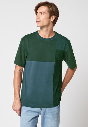 Homme portant un t-shirt à manches courtes bicolore vert foncé et bleu sarcelle avec un logo subtil d'arbre sur la poitrine, assorti à un jean bleu clair.
