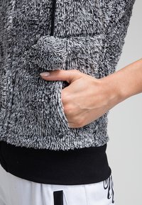 Mīksts pelēks žakets ar samtveida tekstūru, kuram ir rāvējslēdzēja aizdare, melna josta un dekoratīvs pom-pom elements priekšpusē.