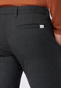 Dunkelgraue maßgeschneiderte Hosen mit einer subtilen Struktur, ausgestattet mit einer Gesäßtasche und einem kleinen Markenetikett. Das Material wirkt leicht und glatt.