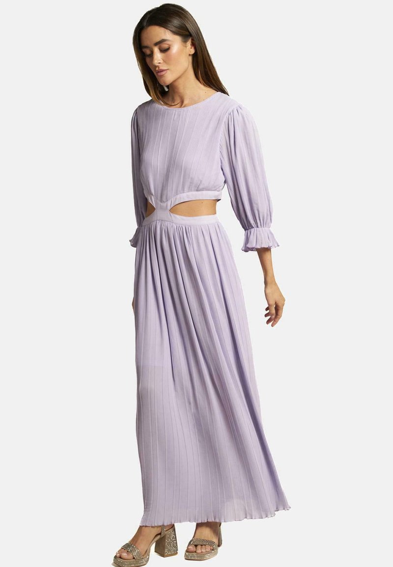 Lila gefaltetes Maxikleid mit langen Ärmeln, Ausschnitt an der Taille, strukturiertem Stoff und Rüschensäumen. Getragen mit metallischen Absatzsandalen.