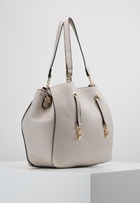 Vaalea harmaa teksturoitu nahkakäsilaukku, jossa on kaksinkertaiset olkahihnat ja kultaiset metalliosat, esillä valkoisella jalustalla.