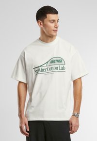 Tricou alb din bumbac, cu mâneci scurte și decolteu rotund. Prezintă un text grafic verde "Another Cotton Lab" și un design cu o rachetă de tenis.