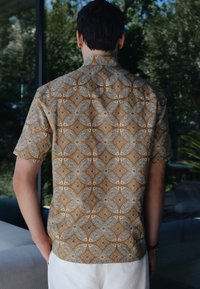 Hombre de pie de espaldas a la cámara, vistiendo una camisa marrón de manga corta con un patrón floral geométrico blanco y pantalones blancos, al aire libre cerca de una ventana de cristal.