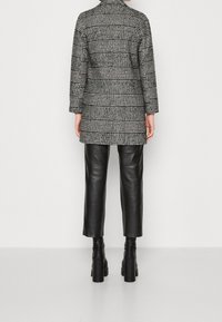 Person in einem schwarz-weißen karierten Mantel, schwarzen Leder-Cropped-Hosen und schwarzen angeleiteten Stiefeletten, der vor einem einfarbigen Hintergrund steht.