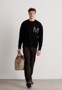 Suéter negro de punto con detalle verde en la "M", combinado con pantalones oscuros. Lleva un bolso de viaje de cuero marrón claro con acentos en contraste.