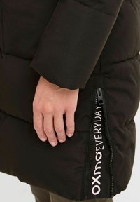 Black puffer jacket featuring a visible zipper with white "OXMO EVERYDAY" text, textured fabric, and a hand resting on the side seam.