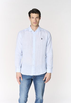 Uomo che indossa una camicia azzurro chiaro a maniche lunghe con logo sul petto e jeans blu, in piedi davanti a uno sfondo chiaro e semplice.