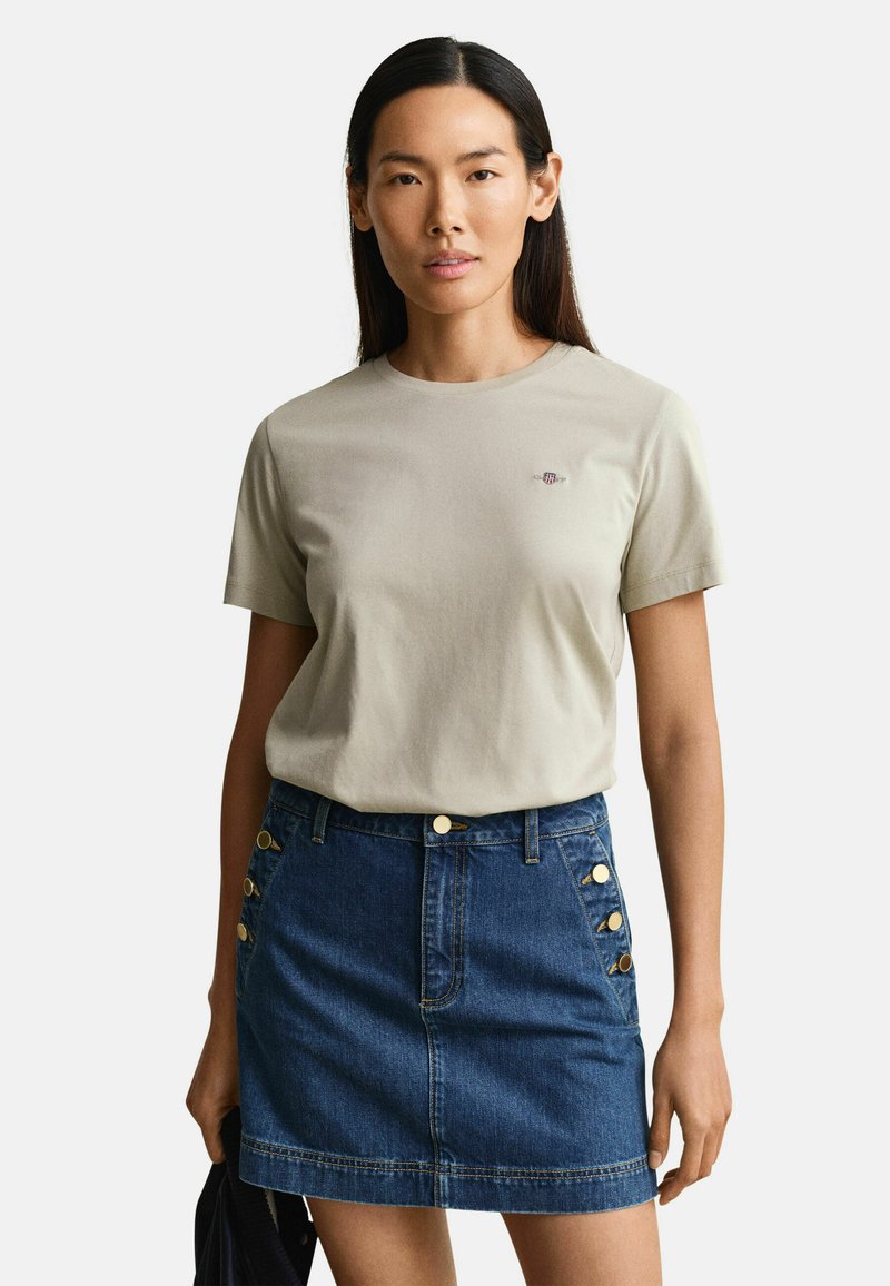 Γυναίκα με μακριά σκούρα μαλλιά, φοράει μια μπεζ μπλούζα με κοντά μανίκια και μια μπλε τζιν φούστα με χρυσά κουμπιά στις τσέπες.