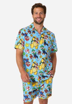 Mann lächelt und trägt ein passendes hellblaues Hemd- und Shorts-Set mit bunten Super Mario Figuren und Symbolen überall.