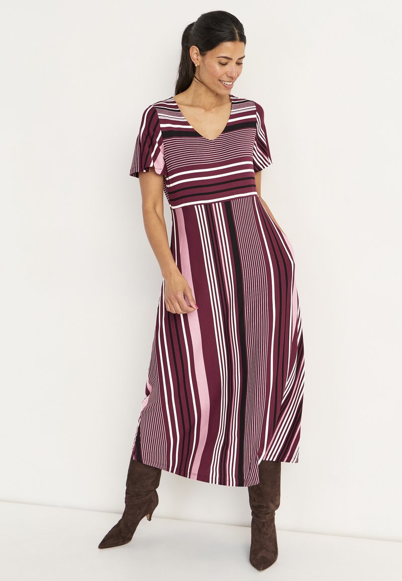 Gestreiftes Maxikleid in Burgunderrot, Rosa und Schwarz; kurze, fließende Ärmel; V-Ausschnitt; mit Seitentaschen; getragen mit knielangen braunen Stiefeln.