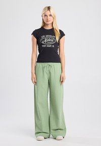 T-shirt noir à manches courtes avec texte imprimé ; pantalon large à carreaux verts avec ceinture élastique ; modèle portant des chaussures blanches décontractées.