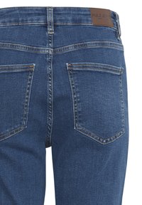 Rückansicht einer blauen Jeans mit zwei Gesäßtaschen, gelber Naht und einem braunen Lederetikett mit Markenlogo am Bund.