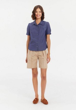Chemise bleu marine à manches courtes avec un col structuré et des boutons, associée à un short beige clair plissé. Le modèle porte des mocassins marron.