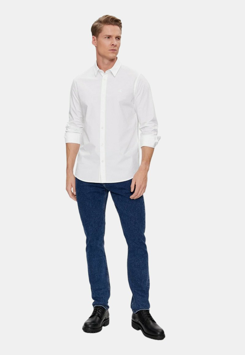 Biała koszula z długim rękawem i zapięciem na guziki, zestawiona z ciemnoniebieskimi dżinsami. Model stoi prosto, nosząc czarne buty, na neutralnym tle.