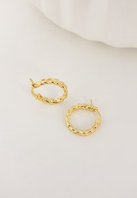 Pendientes de aro de oro con un diseño trenzado, textura suave y cierre de bisagra. Los pendientes son de forma circular.