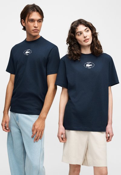 Два модела, облечени в тъмносини тениски с логото на Lacoste, единият с леви светли дънки, а другият с бежови шорти, позиращи пред едноцветен фон.