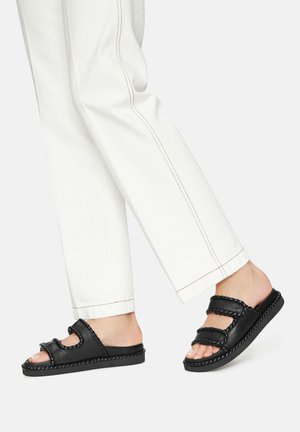 Infradito neri con due cinturini, dettagli intrecciati e suola texturizzata, indossati con pantaloni bianchi a gamba dritta con raffinati cuciture.