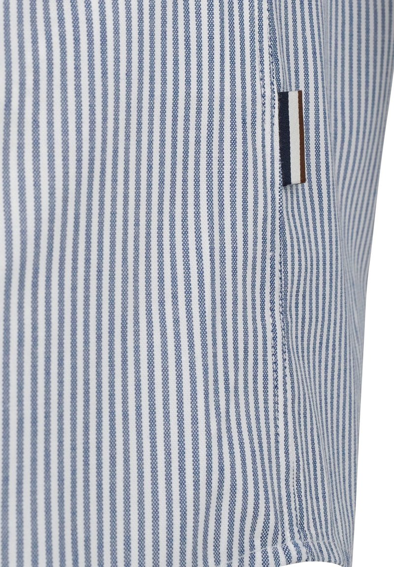Chemise rayée en coton bleu et blanc, présentant des lignes verticales, une étiquette sur la couture latérale avec trois rayures horizontales en marine, marron et crème.