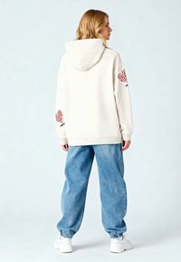 Krämfärgad hoodie med huva, prydd med röda hjärtan på ärmarna och texten "SWEET". Kombineras med lösa blå denimbyxor och vita sneakers.