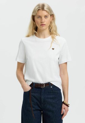 Blonde woman wearing a white T-shirt with a small heart on the chest, dark blue jeans, brown braided belt, and bracelets, standing with one hand in pocket.