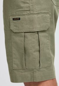 Pantalones cortos de cargo verdes de algodón con bolsillos laterales, costuras visibles y una etiqueta negra "SOLID" en el bolsillo delantero.