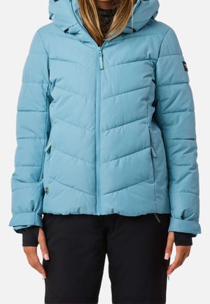 Femme portant une veste d'hiver matelassée bleu clair avec capuche et pantalon noir, debout devant un fond clair uni.