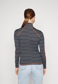 Långärmad, svart mönstrad topp med vita zickzacklinjer, röda detaljer vid sömmarna och design med hög krage. Texturerad, figurnära stil.