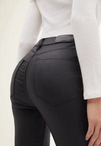 Jeans en denim noir avec une finition brillante, dotés de deux poches arrière, de détails de surpiqûres subtils et d'une étiquette en cuir noir à la taille.