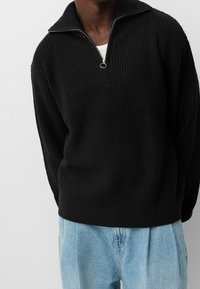 Person som bär en svart ribbad tröja med halvdragkedja över en vit undertröja och löst sittande ljusblå jeans mot en vit bakgrund.