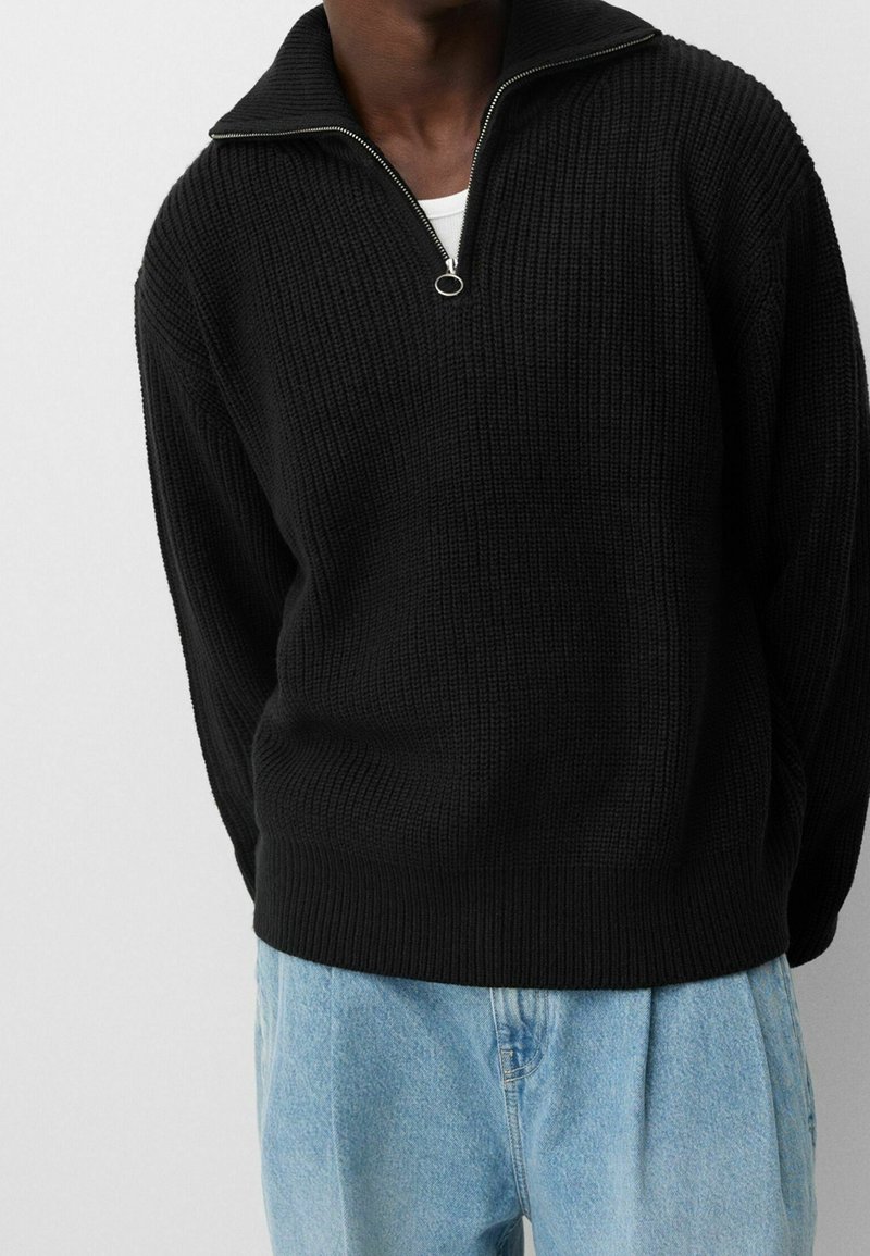 Person som bär en svart ribbad tröja med halvdragkedja över en vit undertröja och löst sittande ljusblå jeans mot en vit bakgrund.