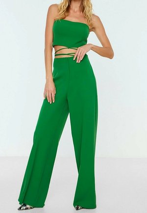 Kobieta ubrana w zielony, bezramiankowy krótki top z wiązaniami do owijania oraz pasujące szerokie spodnie, stojąca na jednolitym białym tle.