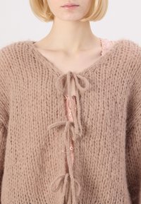 Lys brun strikket cardigan med V-hals, med bånd foran i midten og en myk, teksturert finish. Under er en rosa knappetopp synlig.